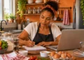 Mulher trabalha de forma autônoma na cozinha, usando notebook e fazendo anotações
