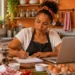 Mulher trabalha de forma autônoma na cozinha, usando notebook e fazendo anotações