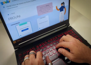 Mãos de pessoa digitando em notebook, tela exibe Portal Prouni gov.br, simbolizando a lista de espera do Prouni.