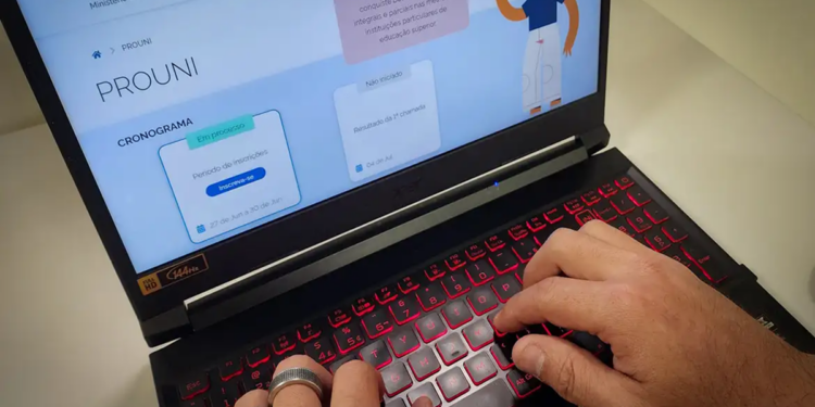Mãos de pessoa digitando em notebook, tela exibe Portal Prouni gov.br, simbolizando a lista de espera do Prouni.