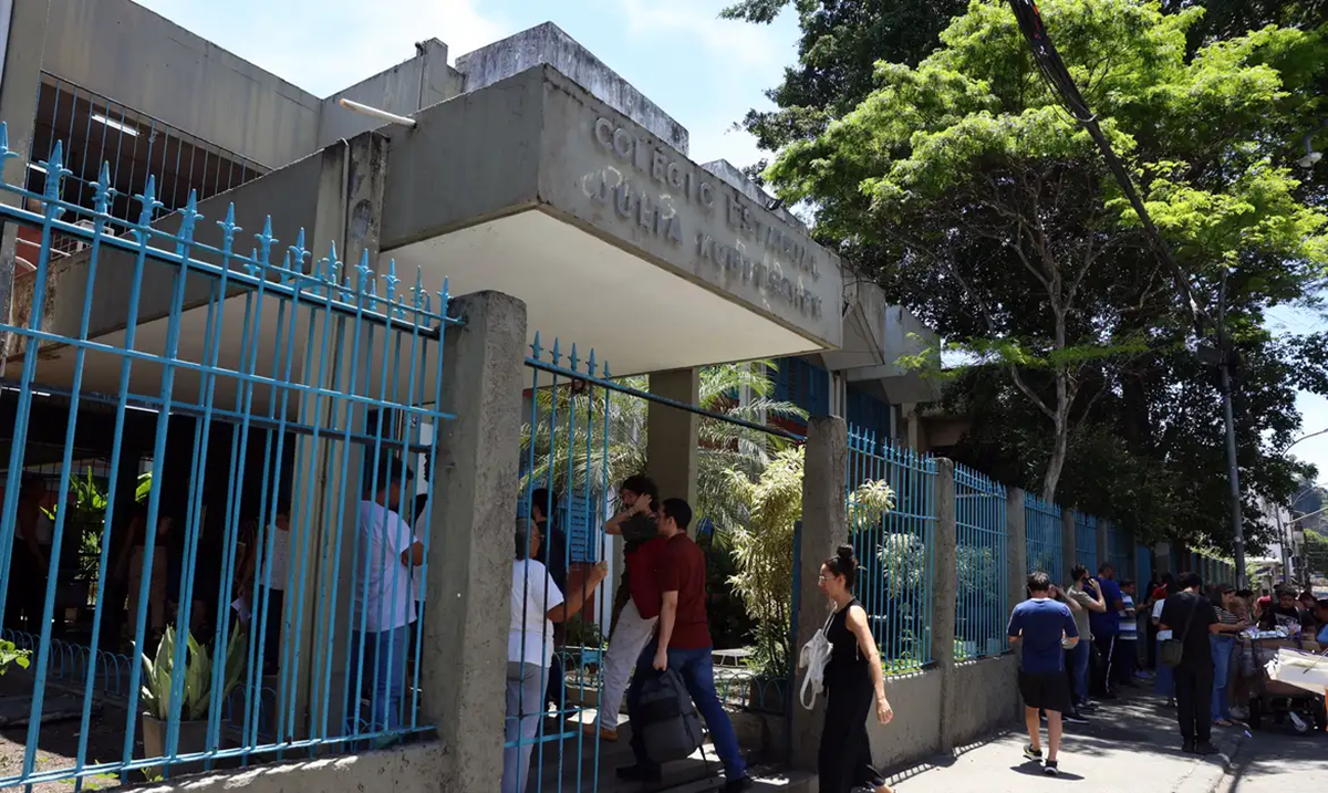Imagem ilustrativa: candidatos entram pelo portão azul de um colégio estadual em dia de exame, cena que remete à Prova Nacional Docente 2026