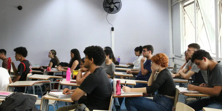 Estudantes sentados em carteiras de uma sala de aula, com cadernos e garrafas de água sobre as mesas, acompanhando a aula durante a preparação para o Provão Paulista.