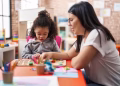 Imagem ilustrativa de profissional de óculos acompanhando uma menina que monta um quebra-cabeça de letras em uma mesa de sala de aula, em referência à psicopedagogia
