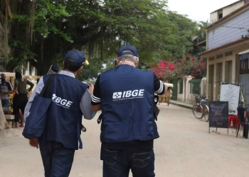 Imagem ilustrativa de dois homens de costas, com coletes e bonés azuis do IBGE, caminhando por uma rua de terra com casas e árvores, em referência à questão do concurso do IBGE