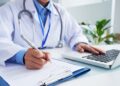 Médico com jaleco azul escrevendo em ficha e utilizando notebook em consultório iluminado, simbolizando o prazo para entrar com recurso no Enamed.