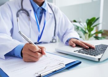Médico com jaleco azul escrevendo em ficha e utilizando notebook em consultório iluminado, simbolizando o prazo para entrar com recurso no Enamed.