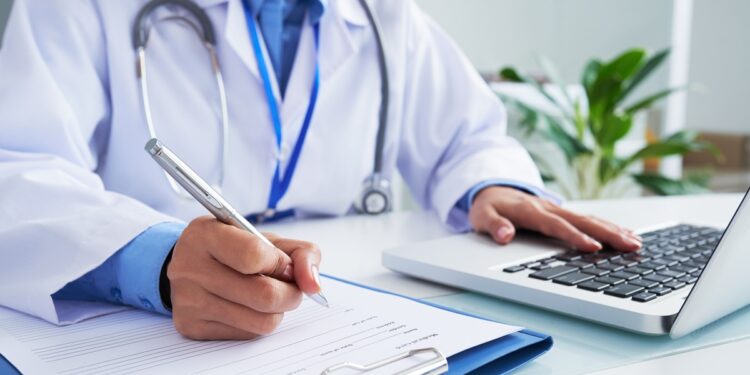 Médico com jaleco azul escrevendo em ficha e utilizando notebook em consultório iluminado, simbolizando o prazo para entrar com recurso no Enamed.