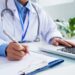Médico com jaleco azul escrevendo em ficha e utilizando notebook em consultório iluminado, simbolizando o prazo para entrar com recurso no Enamed.