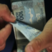 Duas mãos seguram e contam notas de 100 reais em ambiente fechado, simbolizando o pagamento do ressarcimento PIS/Pasep.