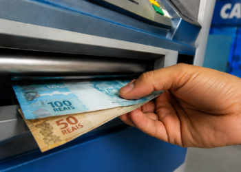 Mão retirando notas de R$ 100 e R$ 50 em caixa eletrônico da Caixa Econômica Federal para representar o pagamento do ressarcimento do PIS/Pasep.