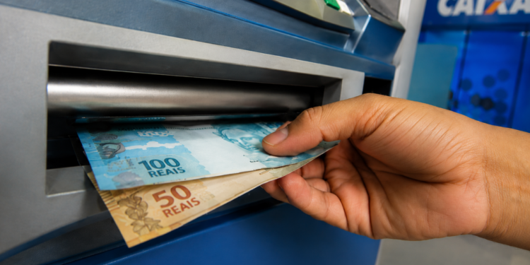 Mão retirando notas de R$ 100 e R$ 50 em caixa eletrônico da Caixa Econômica Federal para representar o pagamento do ressarcimento do PIS/Pasep.
