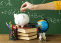 Mão colocando moeda em cofre porco branco sobre livros, globo, moedas e materiais escolares em mesa com quadro verde ao fundo, simbolizando a a diferença de salário entre as etapas da educação básica.