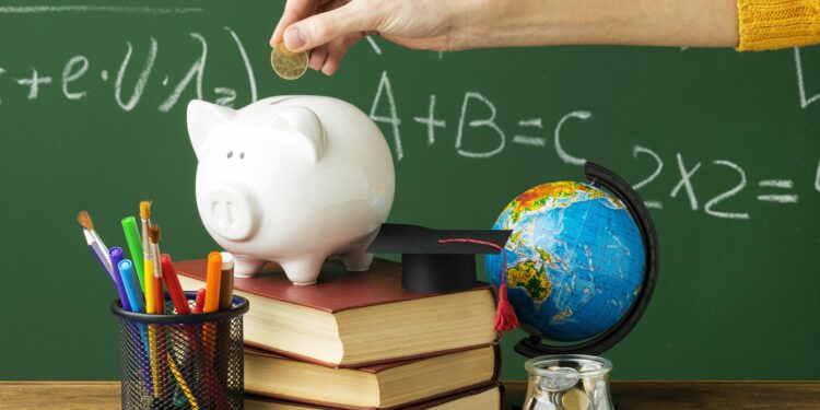 Mão colocando moeda em cofre porco branco sobre livros, globo, moedas e materiais escolares em mesa com quadro verde ao fundo, simbolizando a a diferença de salário entre as etapas da educação básica.