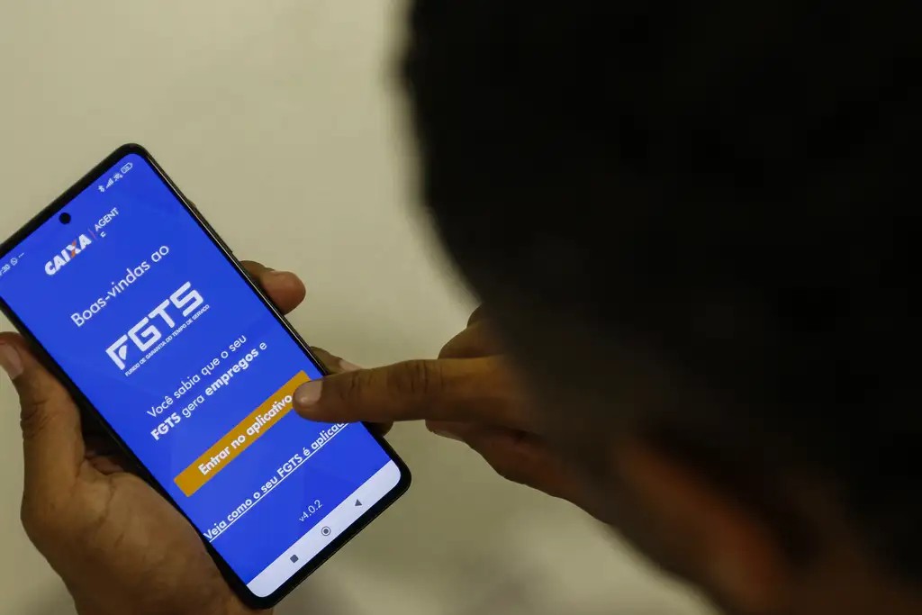Pessoa segurando o celular com a tela de boas-vindas do aplicativo FGTS aberta, tocando no botão de entrada para acessar a conta.