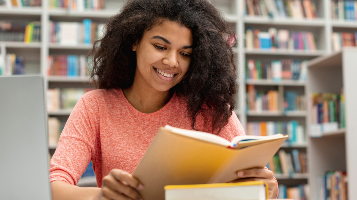 Estudante lê livro em biblioteca durante preparação para questões de Literatura do Enem.