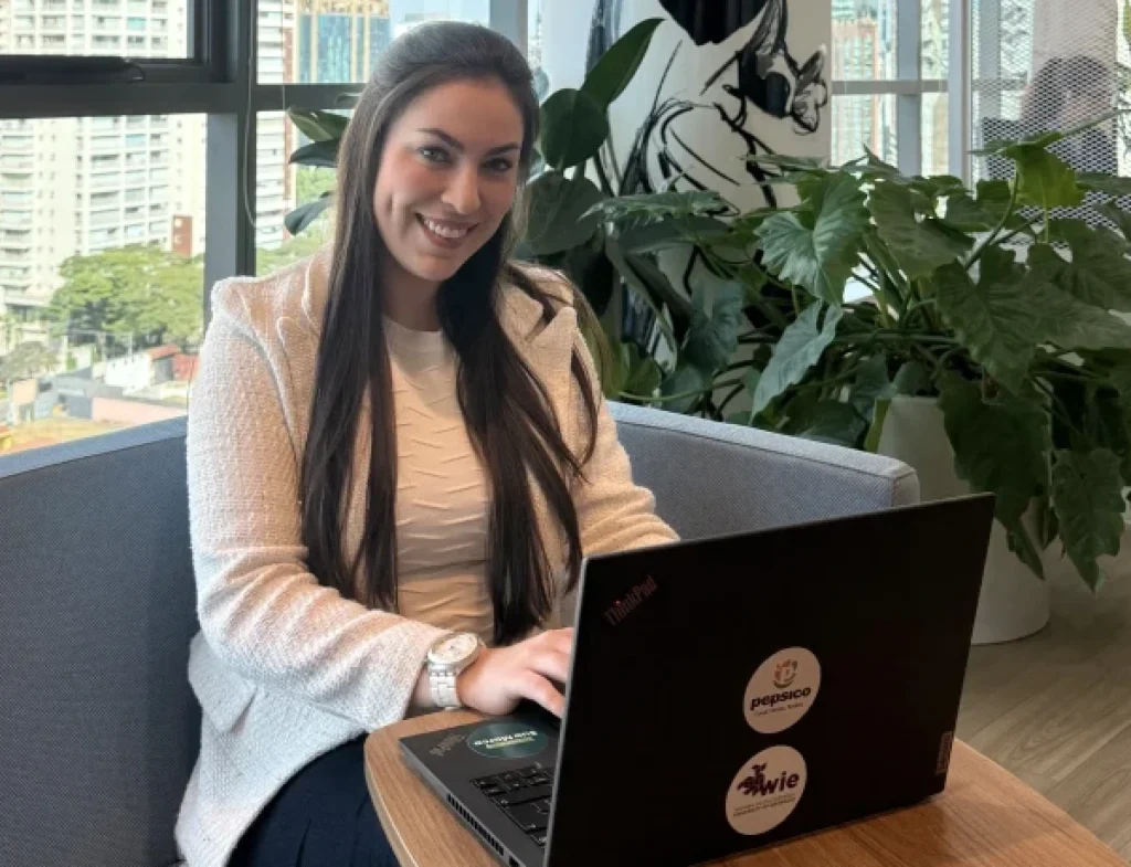 Imagem ilustrativa de profissional sorridente trabalhando em notebook com adesivo da PepsiCo, em escritório com plantas, em referência ao trainee da PepsiCo