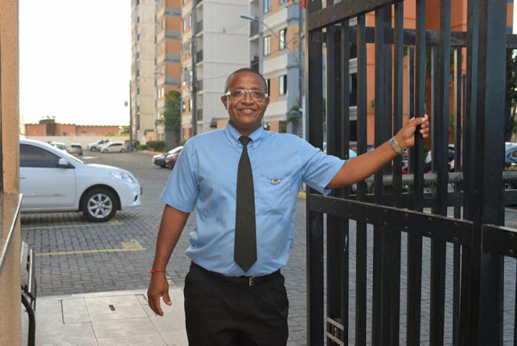 Porteiro uniformizado de camisa social e gravata sorrindo ao lado do portão de entrada de um condomínio residencial.