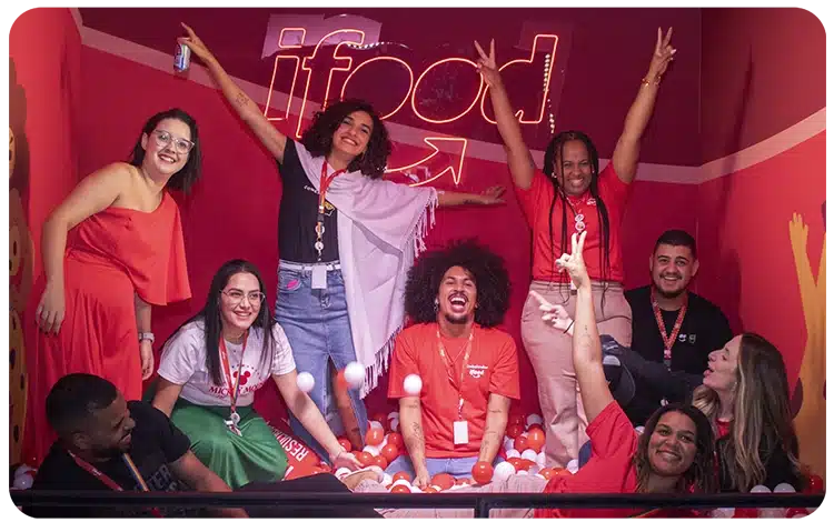 Grupo de profissionais sorrindo e comemorando em ambiente descontraído com a marca iFood ao fundo, representando os times que recebem os participantes do programa jovem aprendiz.