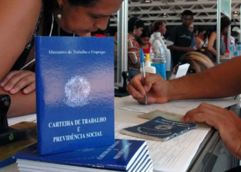 Candidata em balcão de atendimento de um mutirão de emprego com atendente preenchendo formulários, ao lado de exemplares da Carteira de Trabalho e Previdência Social.
