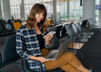 Mulher utilizando notebook e celular em uma área de espera enquanto acessa a internet por Wi-Fi público.