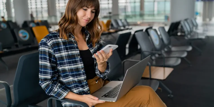 Mulher utilizando notebook e celular em uma área de espera enquanto acessa a internet por Wi-Fi público.