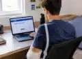 Jovem de costas, com braço engessado, usando notebook no quarto.