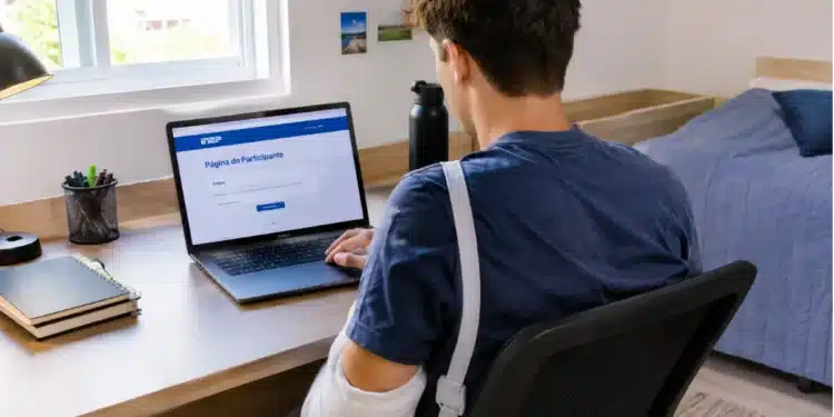 Jovem de costas, com braço engessado, usando notebook no quarto.