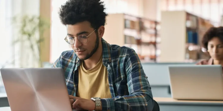 Jovem estudando em biblioteca universitária usando notebook.
