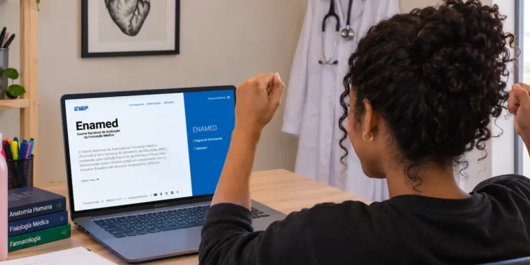 Mulher comemora diante do notebook com a página do Enamed aberta, em um quarto com jaleco e quadro de anatomia.