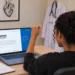 Mulher comemora diante do notebook com a página do Enamed aberta, em um quarto com jaleco e quadro de anatomia.