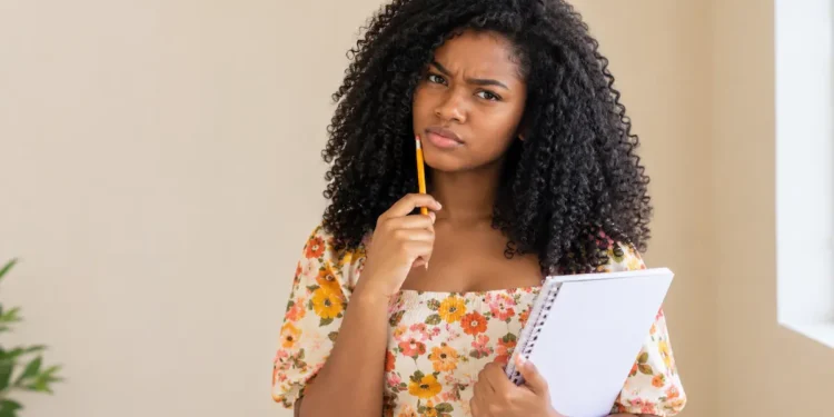 Jovem pensativa segurando caderno com expressão de dúvida em ambiente claro.