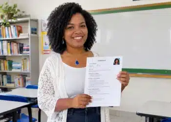 Professora sorrindo e segurando seu currículo em uma sala de aula.