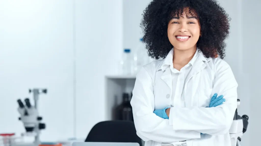 Jovem sorridente usa jaleco branco e luvas azuis em um laboratório.