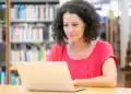 Professora acompanha aula online em biblioteca com livros ao fundo.