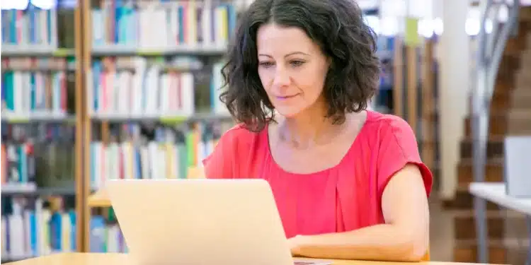 Professora acompanha aula online em biblioteca com livros ao fundo.