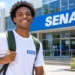 Jovem sorrindo em frente ao SENAI, com blusa branca e mochila nas costas.
