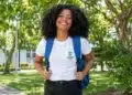 Estudante negra sorrindo, com mochila azul e camiseta do IFS, em um campus arborizado.