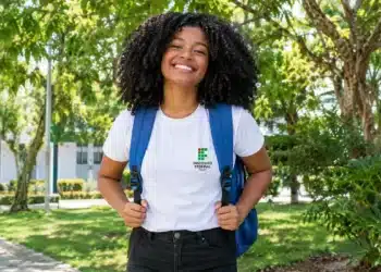 Estudante negra sorrindo, com mochila azul e camiseta do IFS, em um campus arborizado.