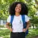 Estudante negra sorrindo, com mochila azul e camiseta do IFS, em um campus arborizado.