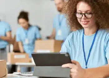 Jovem de cabelo cacheado e óculos usa tablet enquanto outras pessoas organizam caixas ao fundo.