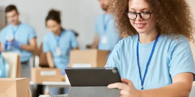 Jovem de cabelo cacheado e óculos usa tablet enquanto outras pessoas organizam caixas ao fundo.