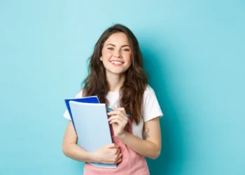 Estudante jovem sorridente com cadernos em fundo azul.