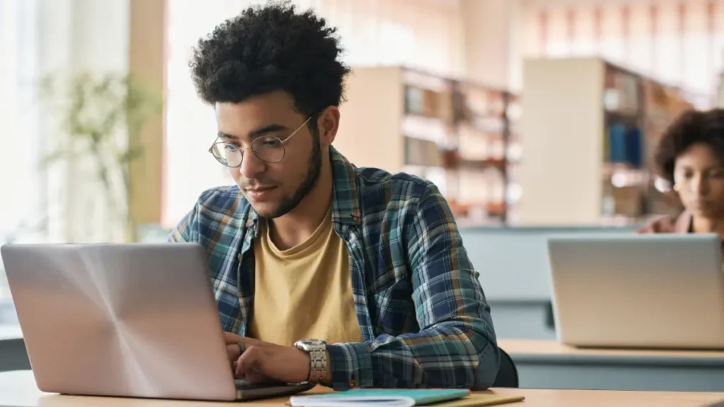 Jovem de óculos e cabelos cacheados usando notebook em biblioteca iluminada.