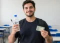 Jovem estudante de camiseta preta e mochila segura uma garrafa de água e um documento de identificação em uma sala de aula com carteiras azuis.