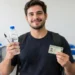 Jovem estudante de camiseta preta e mochila segura uma garrafa de água e um documento de identificação em uma sala de aula com carteiras azuis.