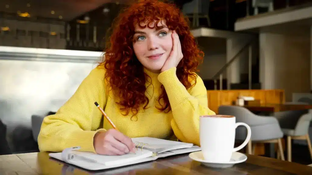Jovem ruiva escrevendo e pensativa em cafeteria moderna.