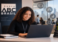 Mulher jovem de cabelos cacheados sorrindo, trabalhando em notebook no Banco ABC Brasil, livros com temas de desenvolvimento e ícones de crescimento ao fundo, simbolizando as inscrições para o estágio Banco ABC Brasil.