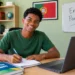 Jovem negro de camiseta verde sorri para a câmera enquanto estuda com notebook e cadernos em um quarto iluminado, com paredes verdes, bandeira de Portugal e quadro escrito “Enem Portugal”.