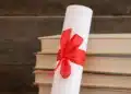 Diploma enrolado com fita vermelha ao lado de livros empilhados.