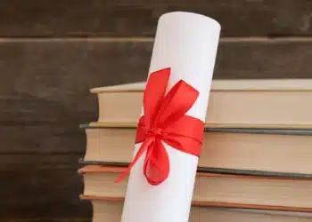 Diploma enrolado com fita vermelha ao lado de livros empilhados.